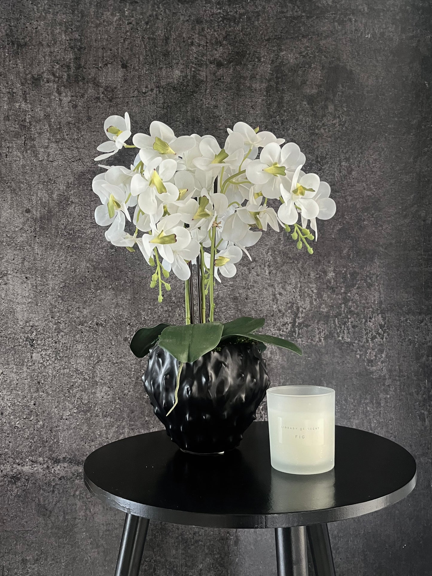 Artificial white orchid surrounded by artificial moss in a contemporary black spiky pot displayed on a table next to a candle.