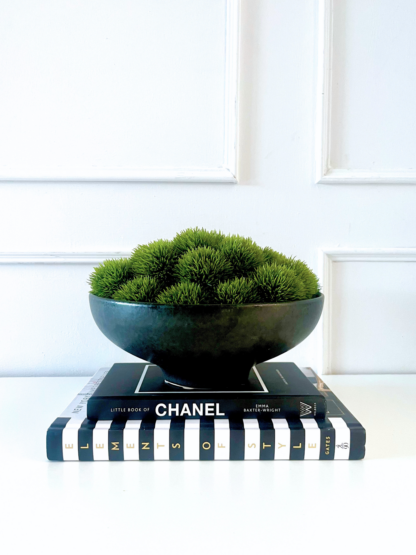 Lush green artificial dianthus set in a black ceramic footed bowl planter displayed on a stack of books.