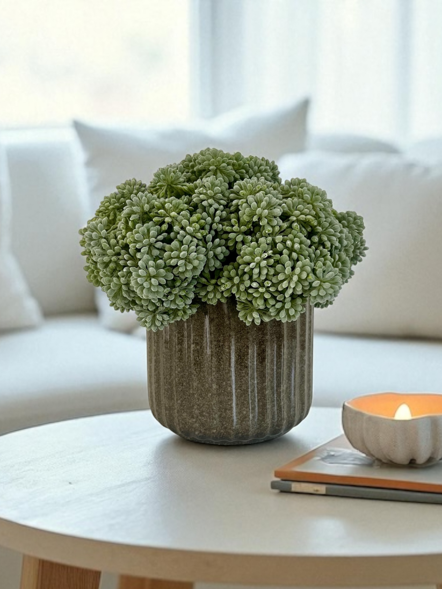 Muted green artificial sedum set in a porcelain ribbed melange green pot displayed on a coffee table next to a lit candle on a stack of books.