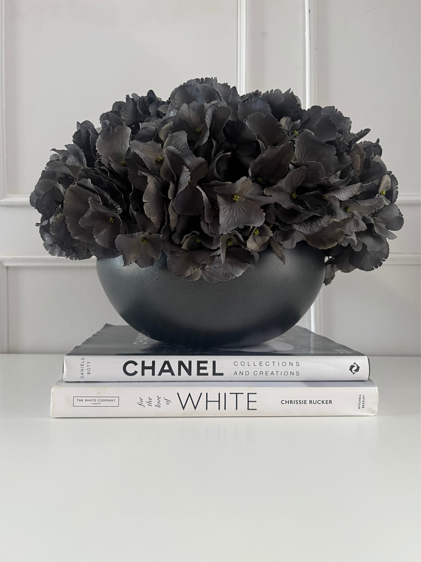 Dark grey artificial hydrangea set in a matt black ceramic bowl planter displayed on a stack of books.
