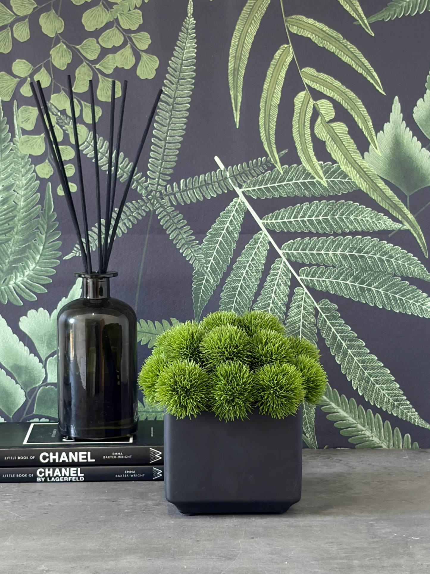 Lush green artificial dianthus set in a black cube planter displayed next to a reed diffuser on 2 books with black and green botanical wallpaper as back drop.