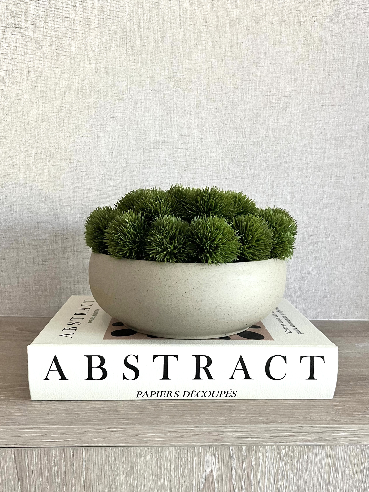 Artificial lush green dianthus nestled in a neutral colour ceramic bowl planter displayed on a book on a wooden surface.