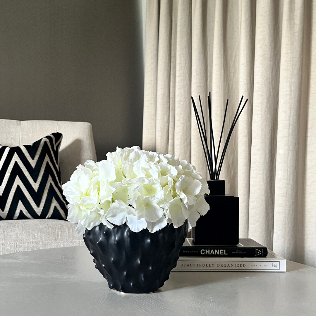 Cream artificial hydrangea set in a spiky black stoneware pot next to a diffuser on a stack of books.