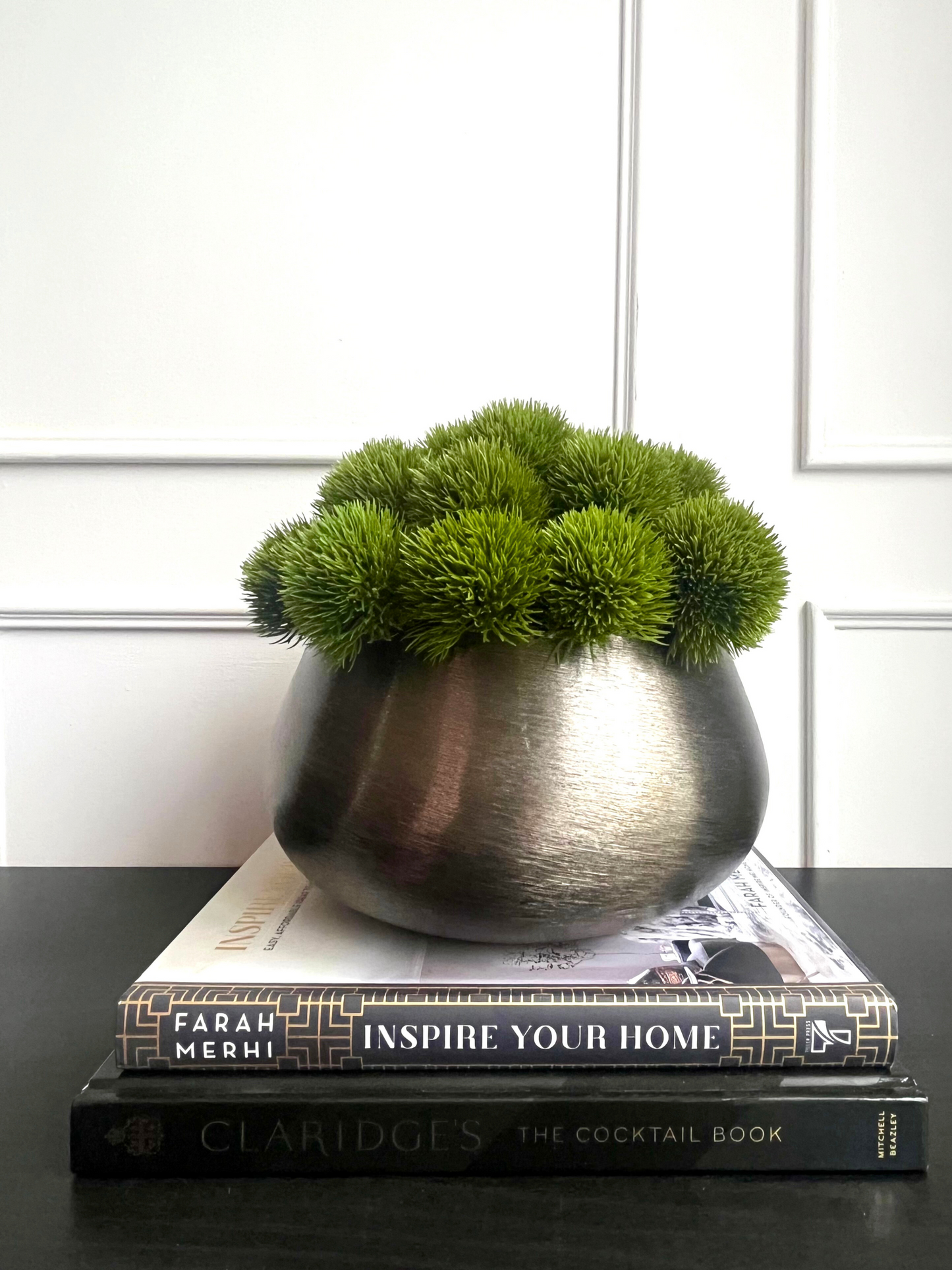 artificial lush green dianthus set in a bronze coloured brush metal planter displayed on a stack of books.