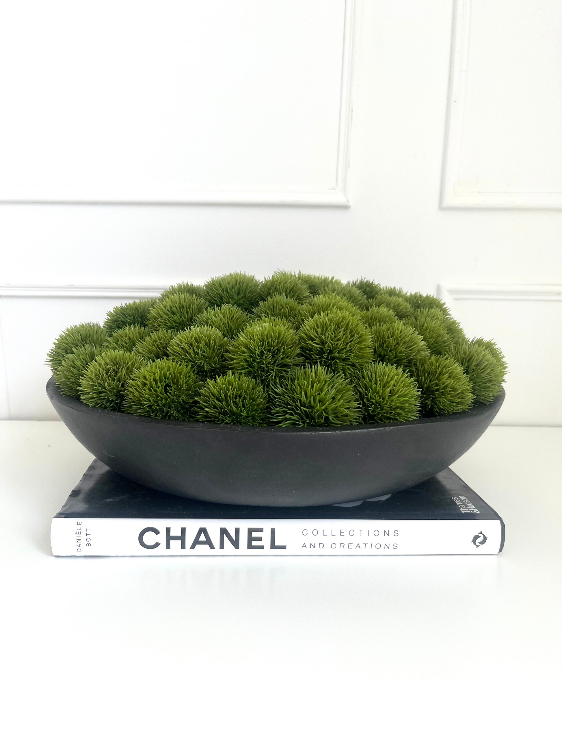 Lush green artificial dianthus set in a large black mango wood bowl displayed on a book.