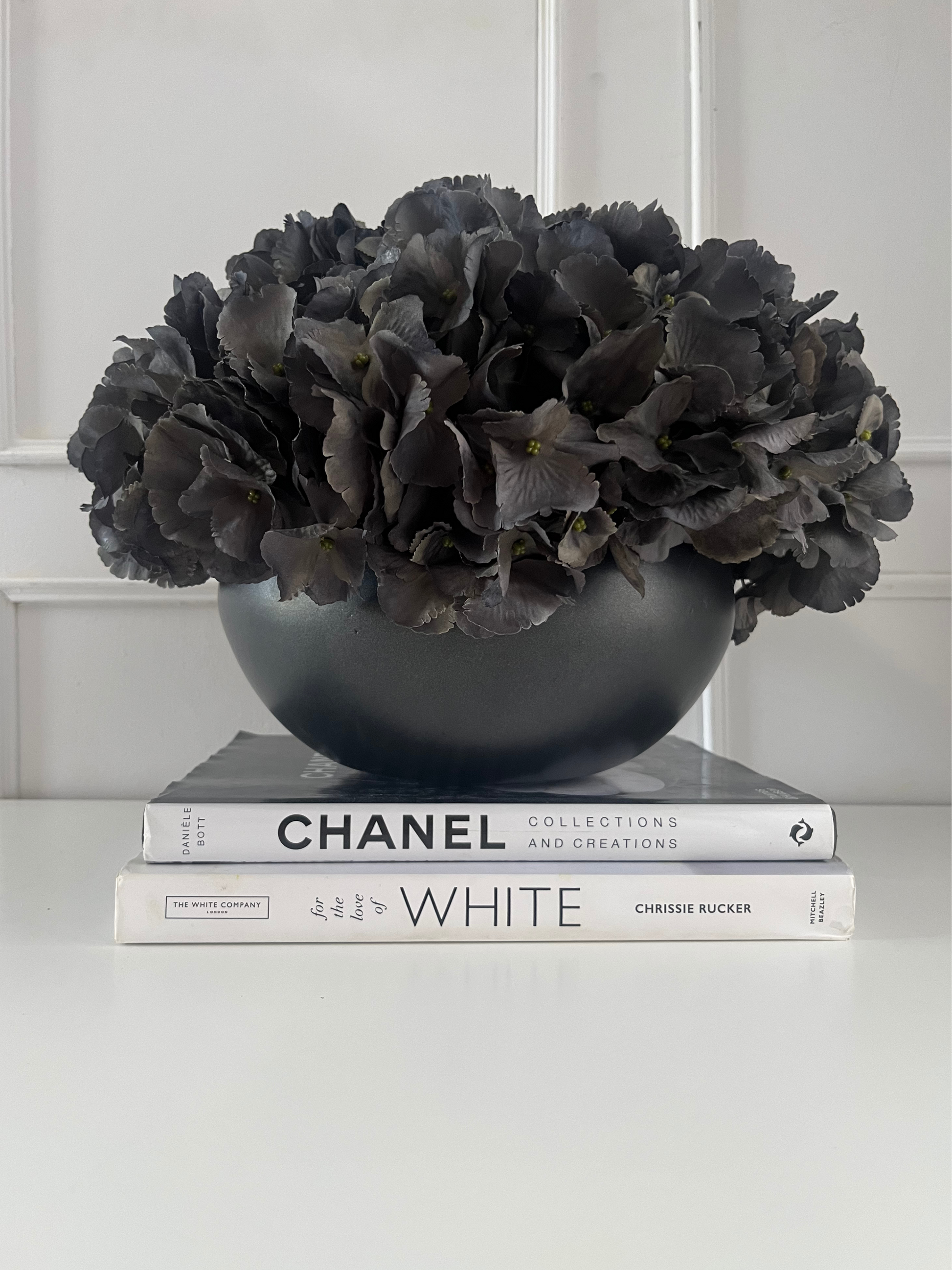 Dark grey artificial hydrangea set in a matt black ceramic bowl planter displayed on a stack of books.