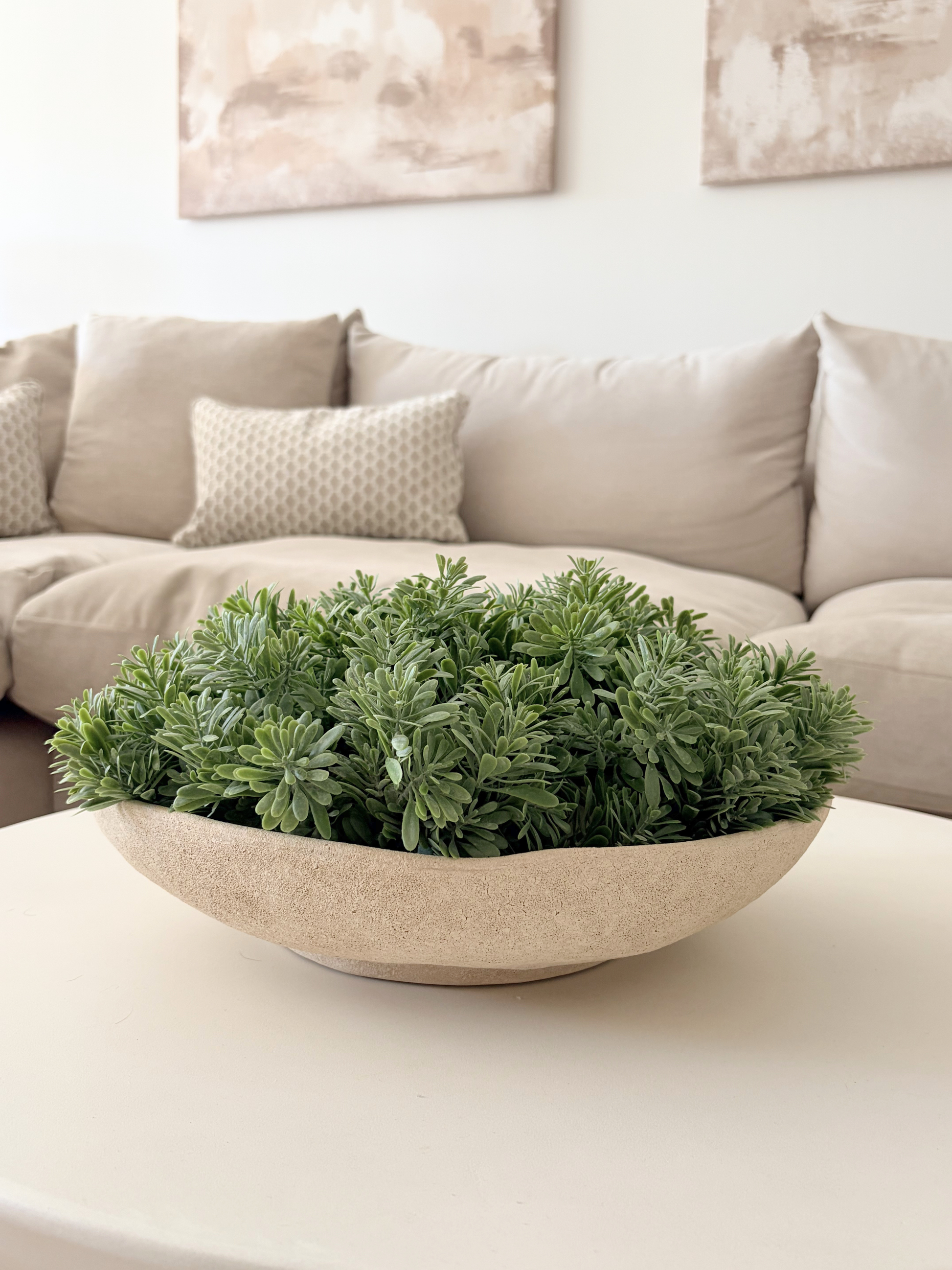 Artificial lush green crossostephium succulents arranged in a stoneware neutral bowl planter displayed on a white coffee table with a neutral sofa and cushions in the background with two canvases on the wall.