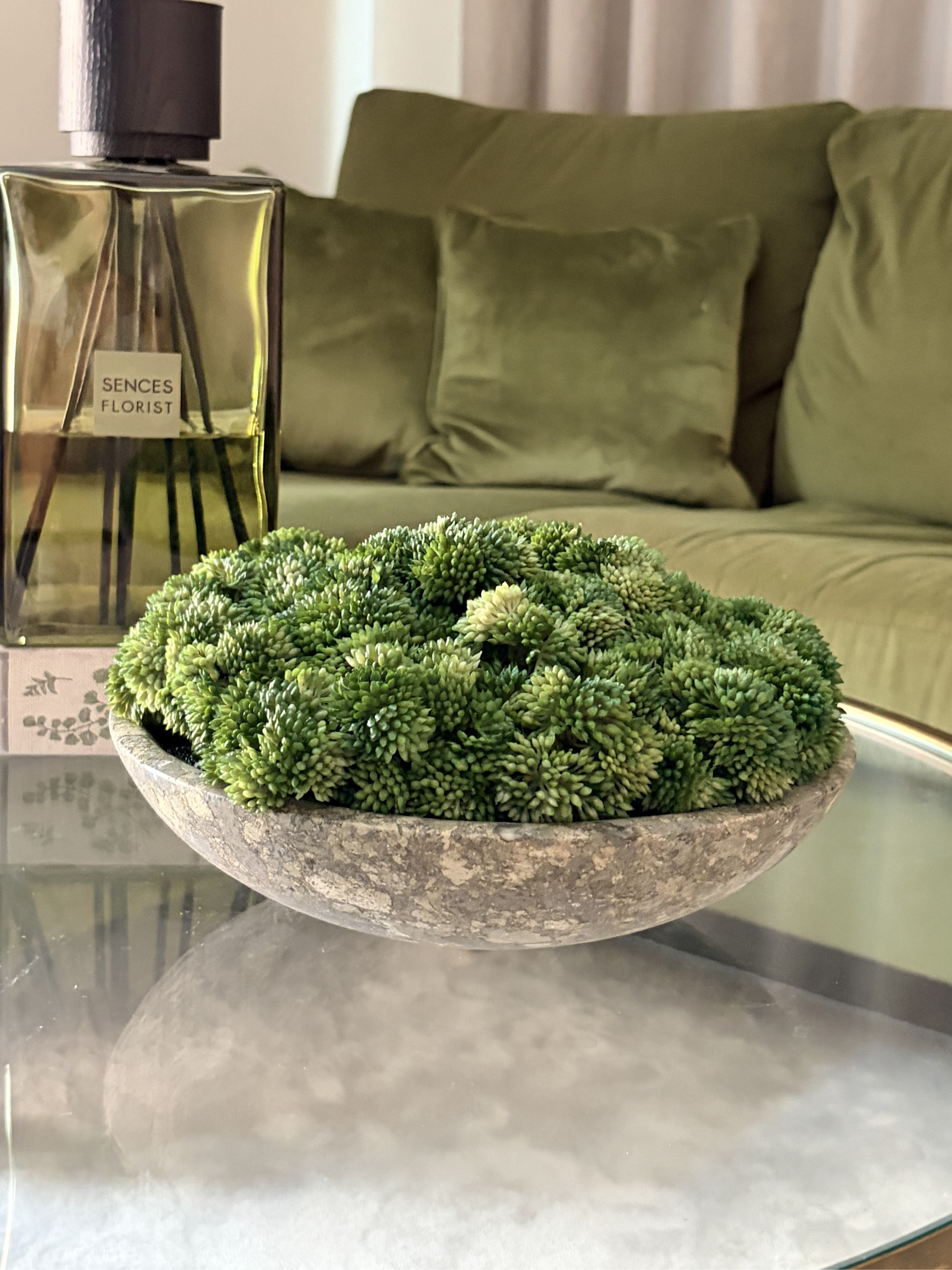 Neutral stoneware bowl brimming with lush green artificial sedum on a glass table next to a green reed diffuser with a green sofa in the background