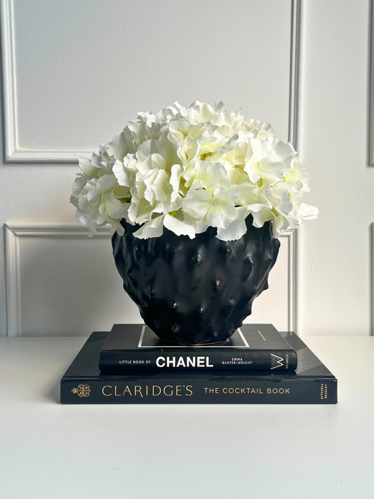 Cream artificial hydrangea set in a spiky black stoneware pot displayed on a stack of books.